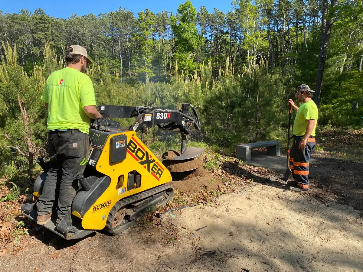 Two crew members augering a planting hole beside a stone bench on a wooded Brewster MA lot